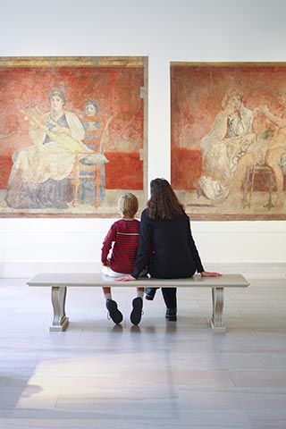 A young boy sits next to his mother on a stone bench looking at a pair of Roman frescoes 