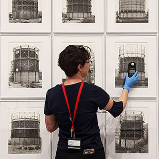 Young woman, facing wall of a grid of framed black and white photographs, holding a small light level measuring instrument up to one photo.