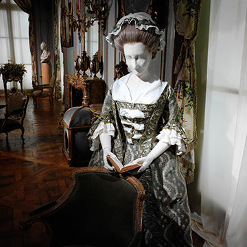 Installation from the Costume Institute’s spring 2004 exhibition “Dangerous Liaisons: Fashion and Furniture in the Eighteenth Century” featuring a female mannequin standing in the Room from the Palais Paar wearing a Robe a la française of green and ivory serpentine silk damask with matching fly-fringe trim and reading a book.