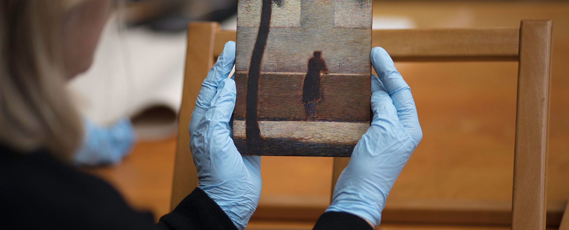 View from over the shoulder of a conservator holding a small rectangular painting in blue rubber gloves; the painting is rendered in short brush strokes that form a kenetic image of a dark, silhouetted tree trunk in the foreground and lone silhouetted figure standing and leaning against a solid, waist-high wall overlooking a faint building in the distance.