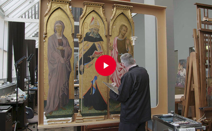 A wide shot of a paintings conservator working on three panels from an Italian altarpiece depicting three religious figures on a gold background. A white triangular play-button set with a red circle appears atop the center of the full image.