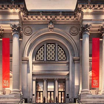 The facade of a building composed of three triumphal arches joined by huge pairs of columns with a very wide tiered staircase leading up to the center arch. Two red Met-branded banners frame the center arch.