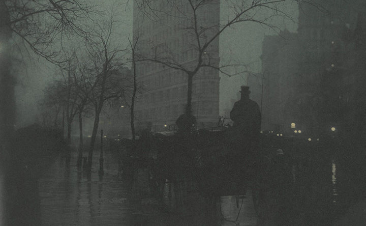 A dark misty scene of people walking on a tree lined sidewalk in New York City, the Flatiron building visible in the background.