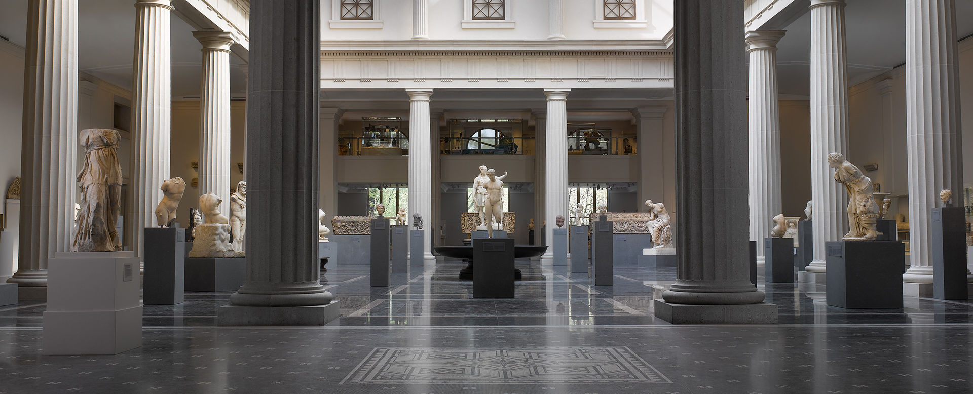 A very large gallery enclosed by a colonnade and filled with with stone sculptures from ancient Rome lit by natural sunlight.