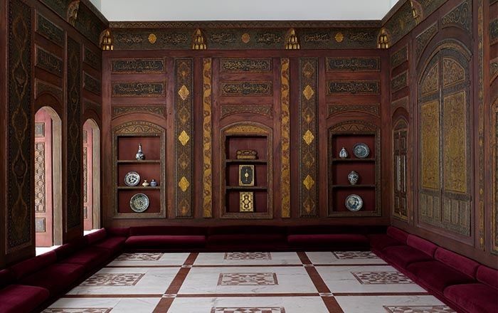 A room from Damascus decorated with gilded dark wood paneling, low dark red velvet covered cushions, and a red and white stone floor 