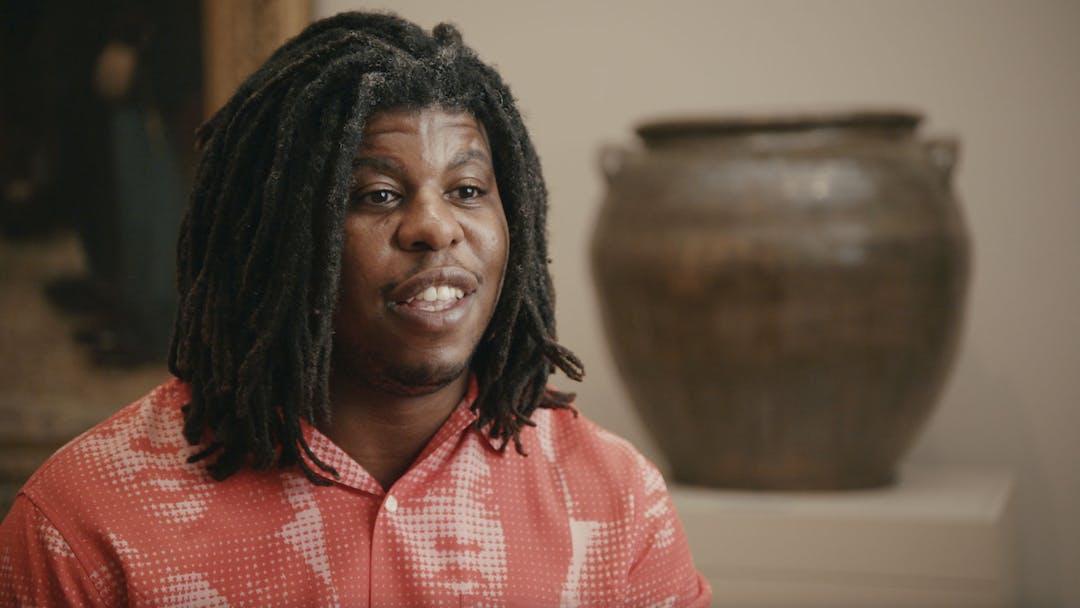 Artist Jon Gray, a dark-skinned male with medium-length braids, sits smiling during an interview in front of a pottery piece presented at the Hear Me Now: The Black Potters of Old Edgefield, South Carolina exhibition.