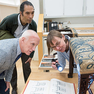 A conservator holding a flashlight and two men looking closely at an upholstered chair and a book showing similar chair designs