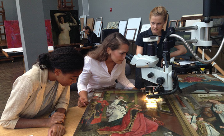 Conservator Sophie Scully and two curatorial fellows gather around a table in the Paintings Conservation studio. One of the fellows looks at a Flemish painting through a microscope while the others observe.