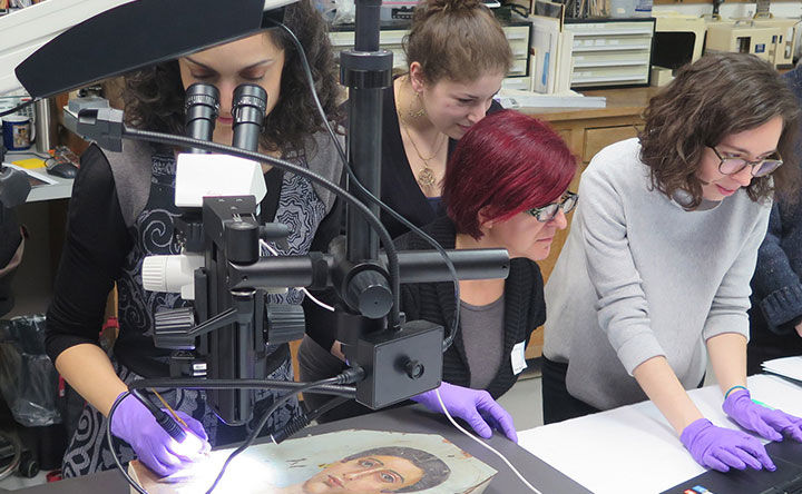 A group of scientists gather around a painting laying flat on a table under a microscope. One person looks at the painting through the microscope lens while three others look out towards what's probably a computer monitor.