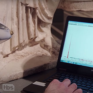 A close of view of a scientist using a laptop to read imaging data collected from a sculptural artwork visible in the background