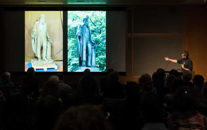 A silhouetted seated audience looks out towards a spotlit speaker presenting in front of large projection screen showing two images of a sculpture side-by-side