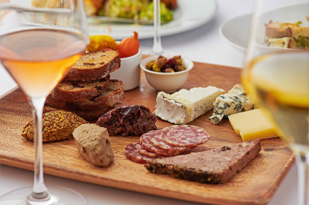 A wooden cheese board filled with an assortment of cheese, deli meats, and bread, are photographed between two glasses of white wine against a white background. 