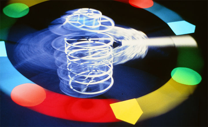A still from Erika Beckman’s digital video called “You the Better.” The still depicts a coil shaped cylinder that emits a cool white and blurred light and seems to spin with centrifugal force. A flat and wide band of alternating ice blue, neon red, yellow, and green bands form a ring around the central coil.