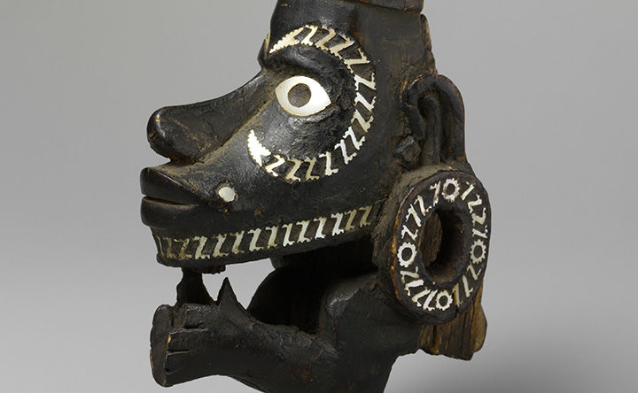 Painted wooden canoe figurehead wearing circular ear ornaments and with small arms with hands raised to the chin.